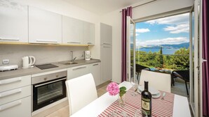 A modern kitchen with a built-in oven, a coffee maker, and a kettle. There is a dining area with a table set for two, a bottle of wine, and a vase with flowers. The room has a large window with a view of a lake and mountains. - Amazing apartment in Cavtat with house sea view (Cavtat)