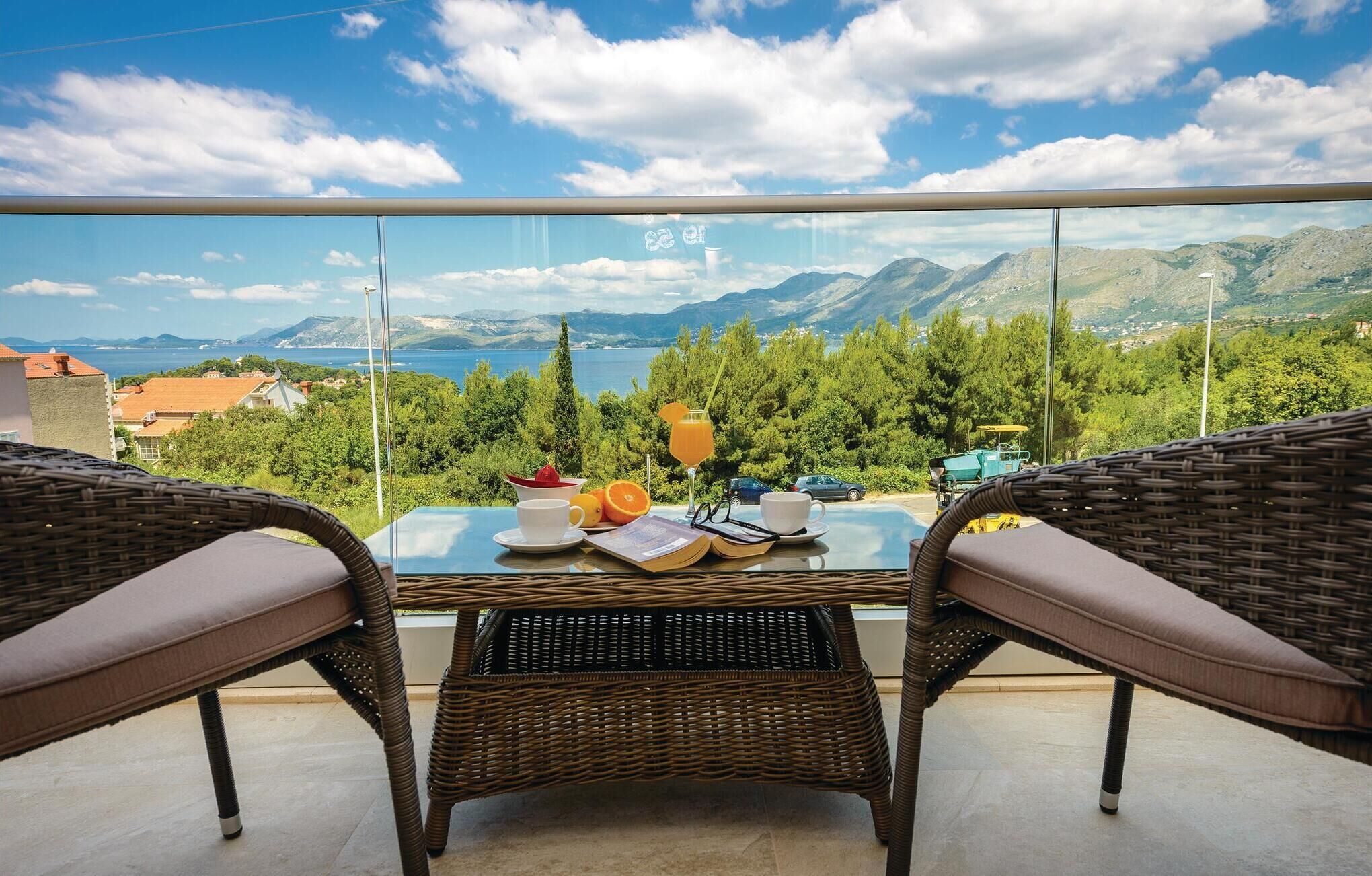 Varanda com móveis de vime, mesa com um livro, copos e um copo de suco de laranja, com vista para uma paisagem montanhosa e um corpo de água.