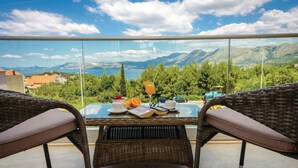 A balcony with wicker furniture, a table with a book, glasses, and a glass of orange juice, overlooking a mountainous landscape and a body of water. - Amazing apartment in Cavtat with house sea view (Cavtat)