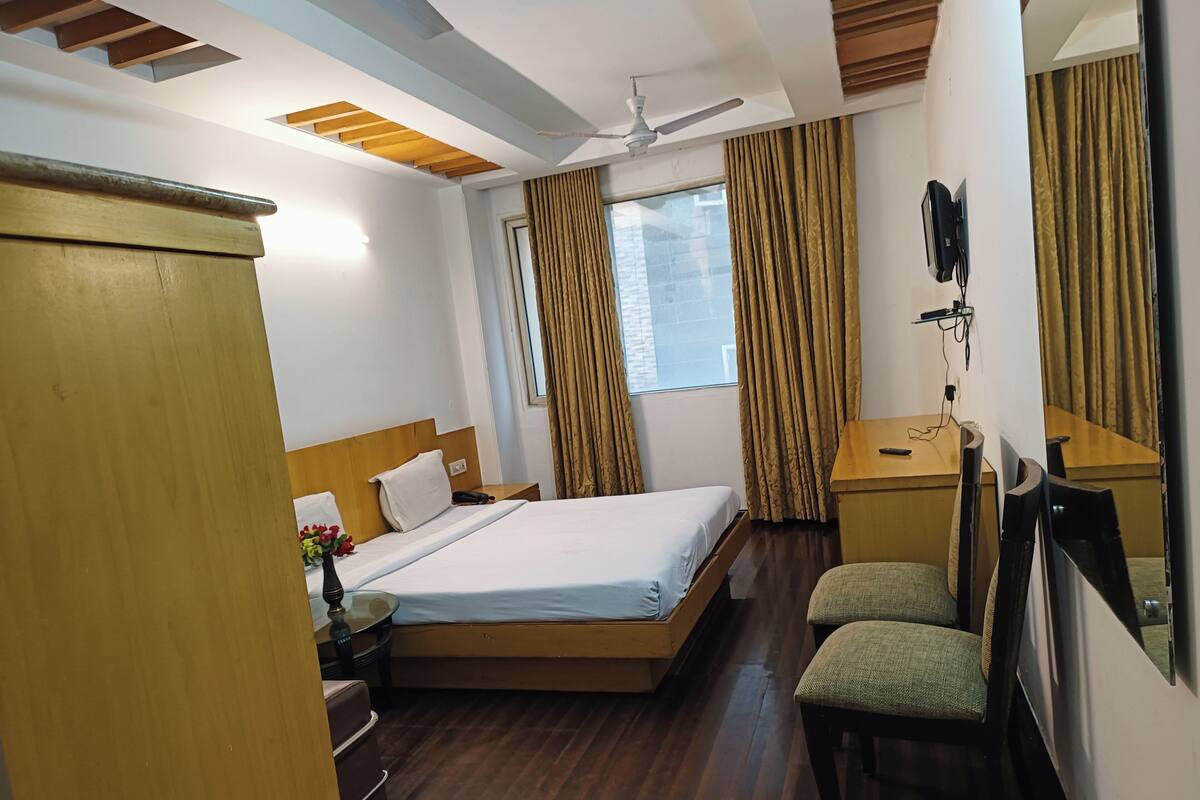 A hotel room with a bed, a desk, a chair, a wardrobe, a ceiling fan, and a window with curtains.