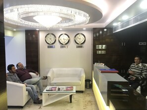 A reception area with a white sofa, a reception desk, and three clocks displaying times from USA, India, and London. - Hotel Gauranga Nearest New Delhi Railway Station (New Delhi)