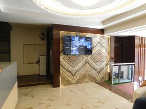 A lobby with a reception desk, a mounted TV displaying multiple screens, a patterned wall, and a glass partition. - Hotel Gauranga Nearest New Delhi Railway Station (New Delhi)