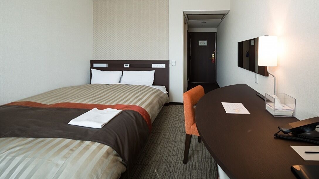 A hotel room with a bed, a desk, a chair, a lamp, and a door.