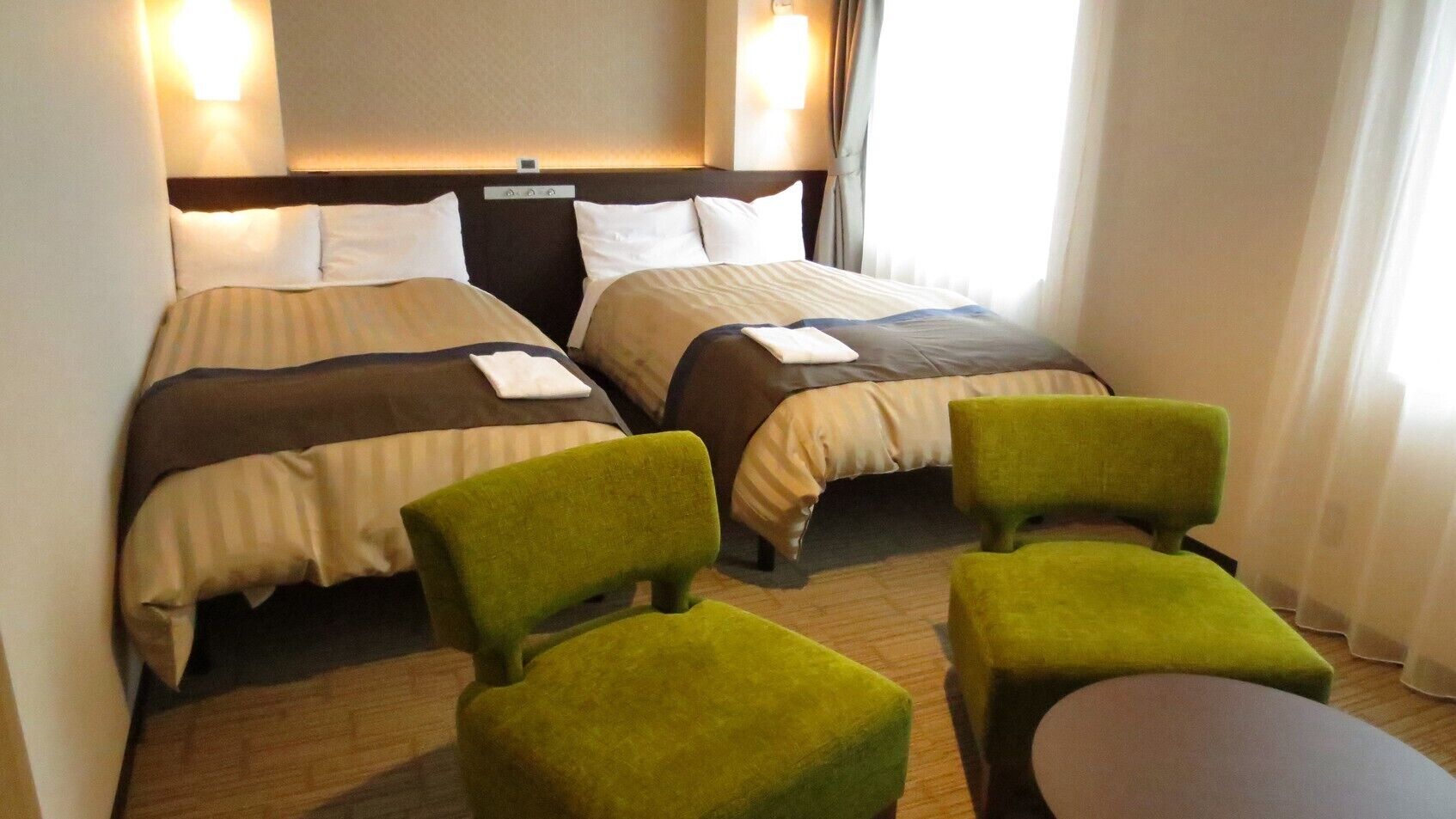 A hotel room with two beds, a headboard, a small table, and two green armchairs.
