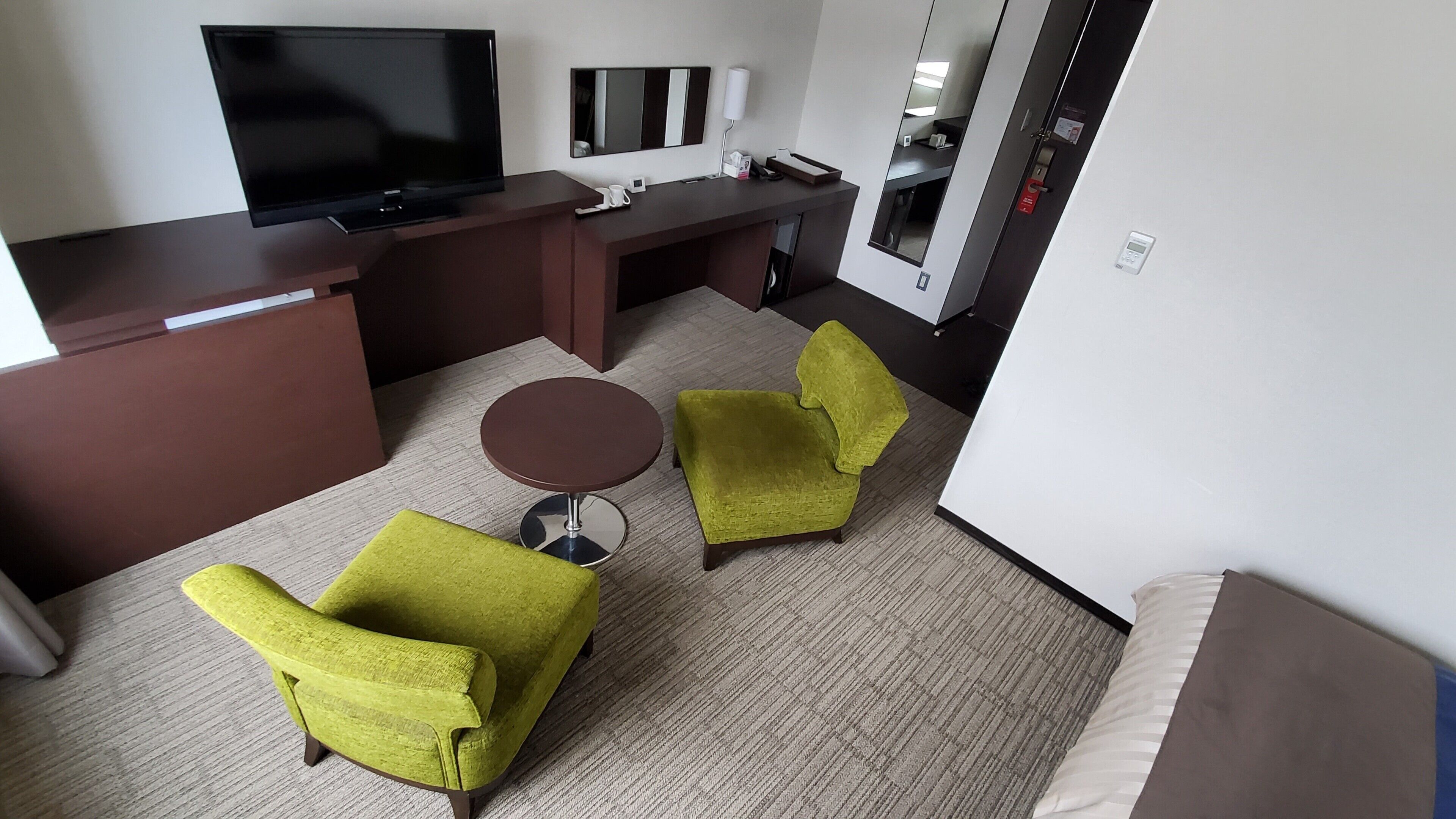 A hotel room with a flat-screen TV, a desk, a chair, a smaller chair, and a round table.