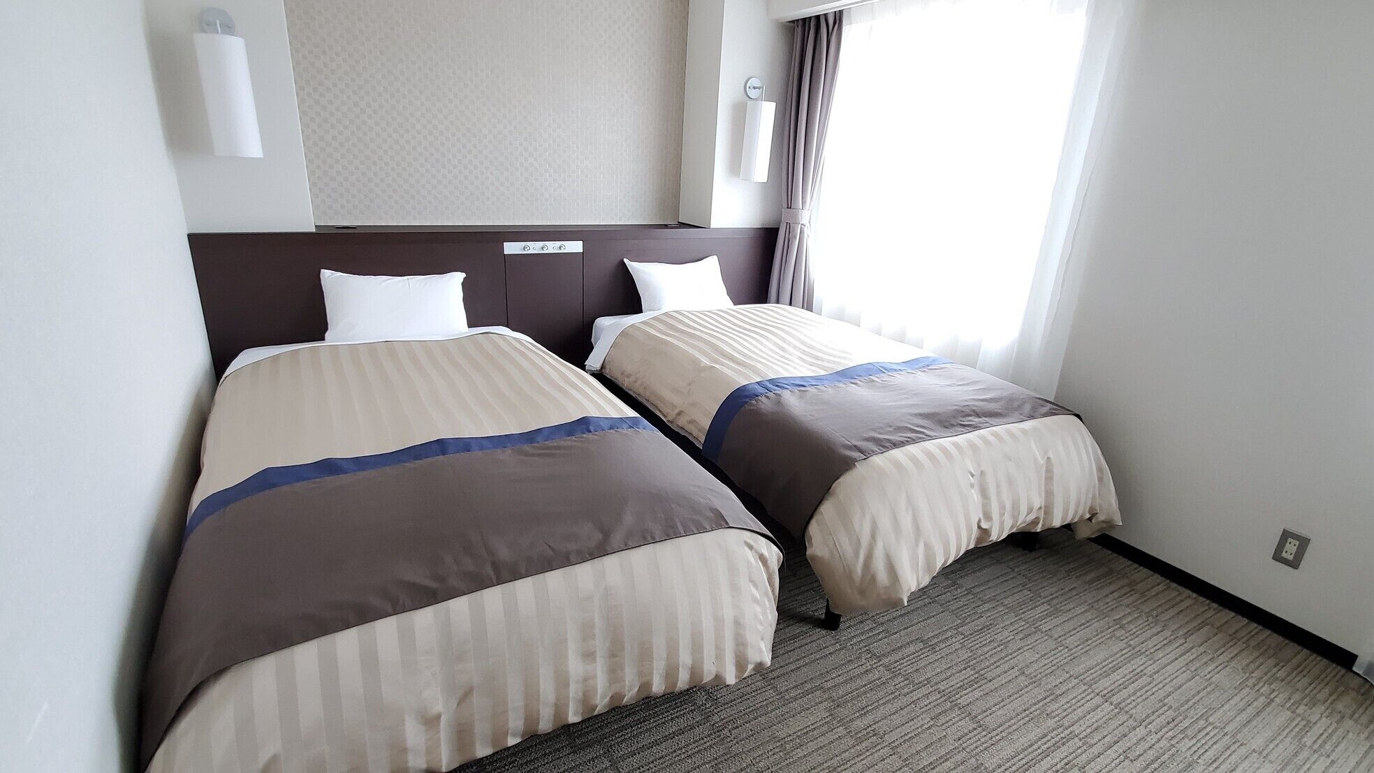A hotel room with two beds, a headboard, a wall-mounted light, a window with curtains, and a carpeted floor.