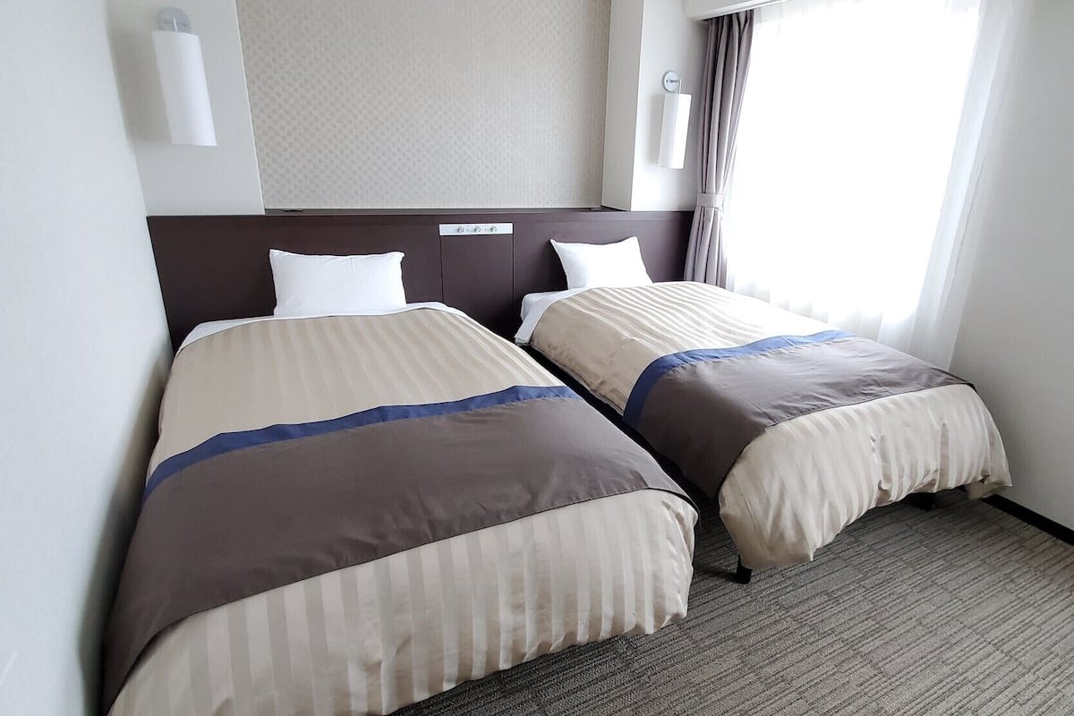 Ein Hotelzimmer mit zwei Betten, einem Kopfteil, einer Wandleuchte, einem Fenster mit Vorhängen und einem Teppichboden.