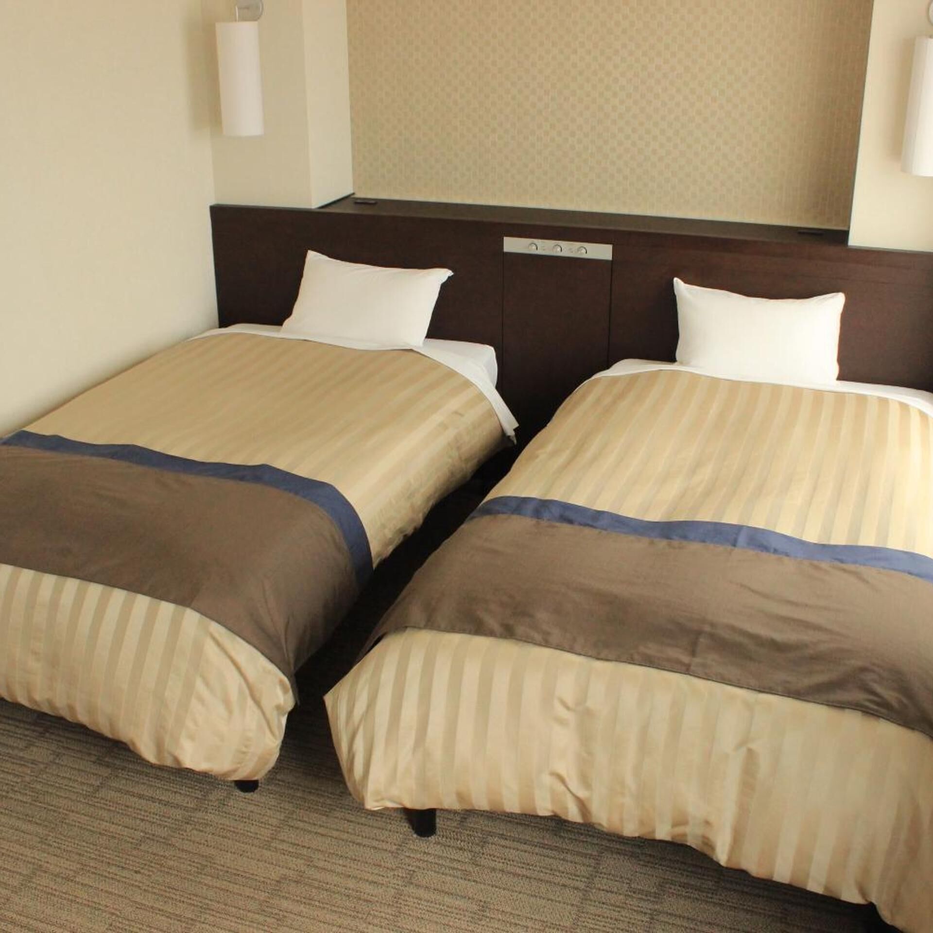 A hotel room with two beds, a headboard with controls, and a carpeted floor.