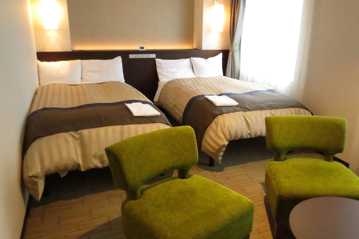 Ein Hotelzimmer mit zwei Betten, einem Nachttisch, einer Lampe und zwei grünen Stühlen.