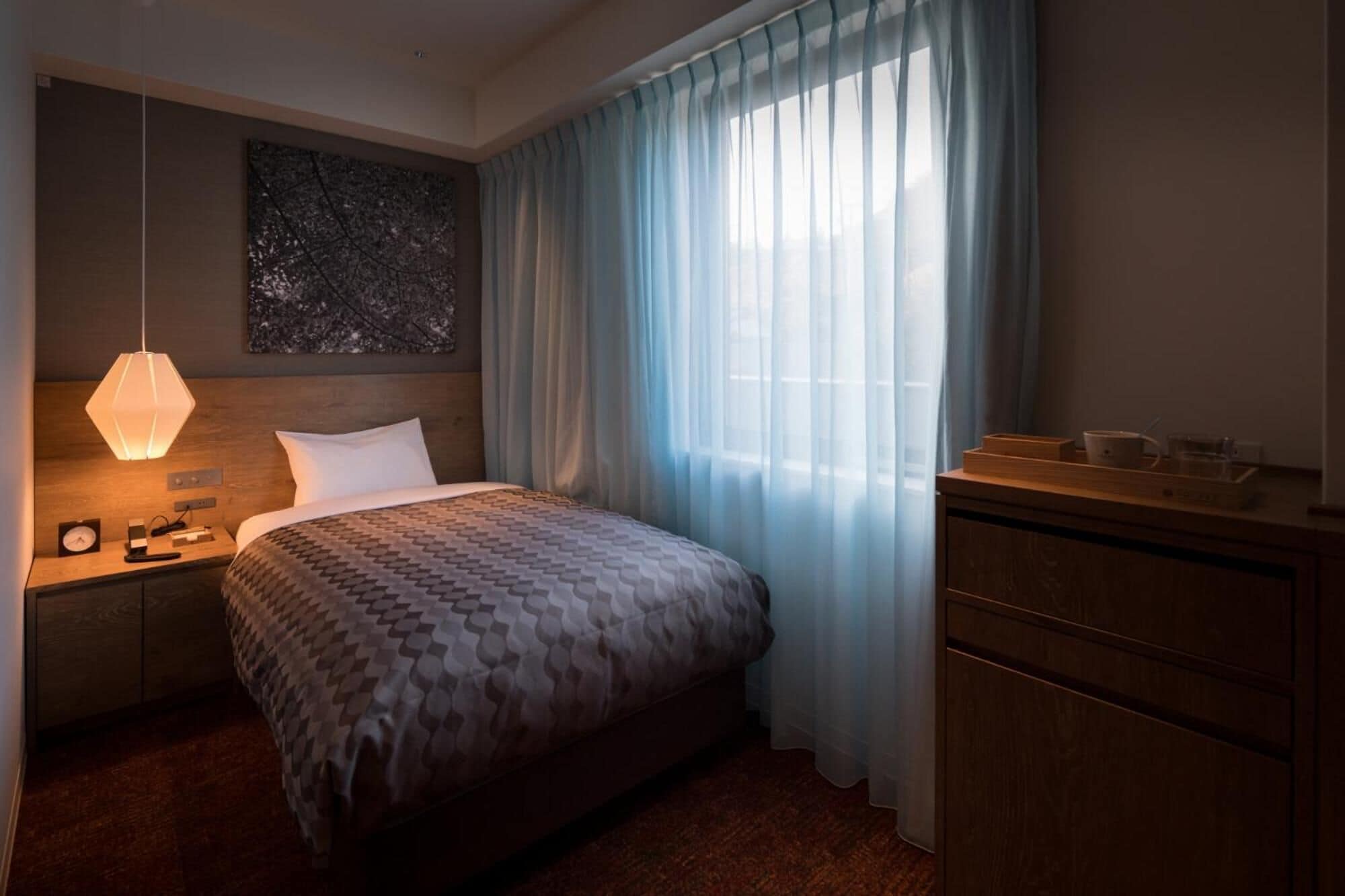 Quarto de hotel com cama, mesa de cabeceira com abajur, cômoda e uma janela grande com cortinas leves.