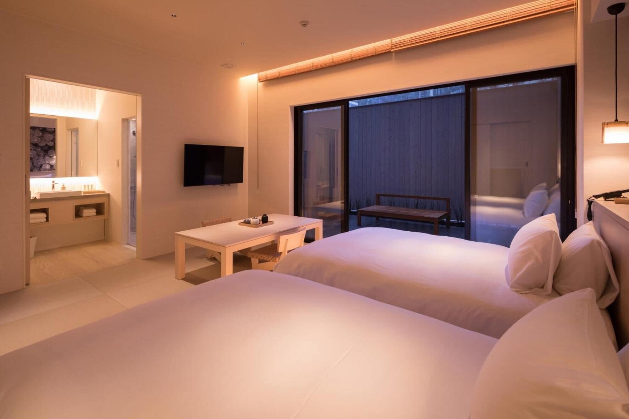 A hotel room with two beds, a small table, a TV, and a sliding glass door leading to a balcony.