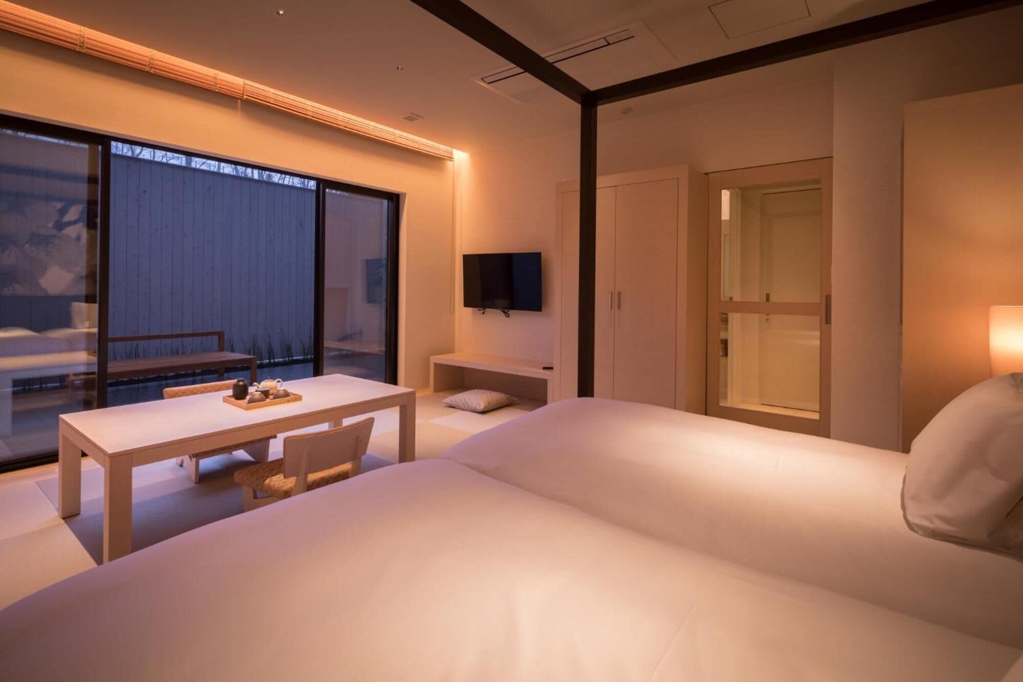 A modern hotel room with a large bed, a small table with tea set, a TV on the wall, and a sliding glass door to a balcony.