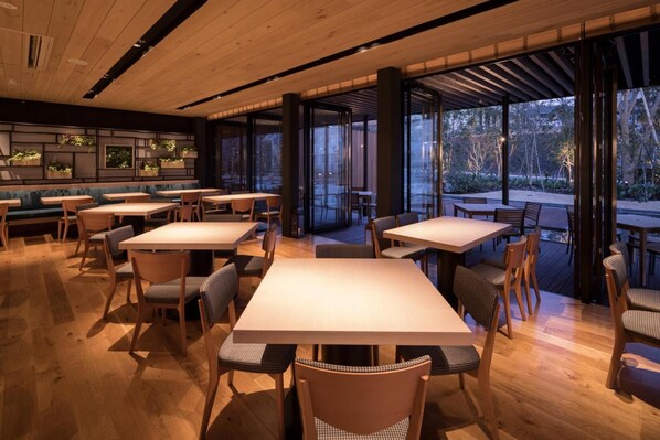 A restaurant with wooden tables and chairs, large windows, and a view of trees.
