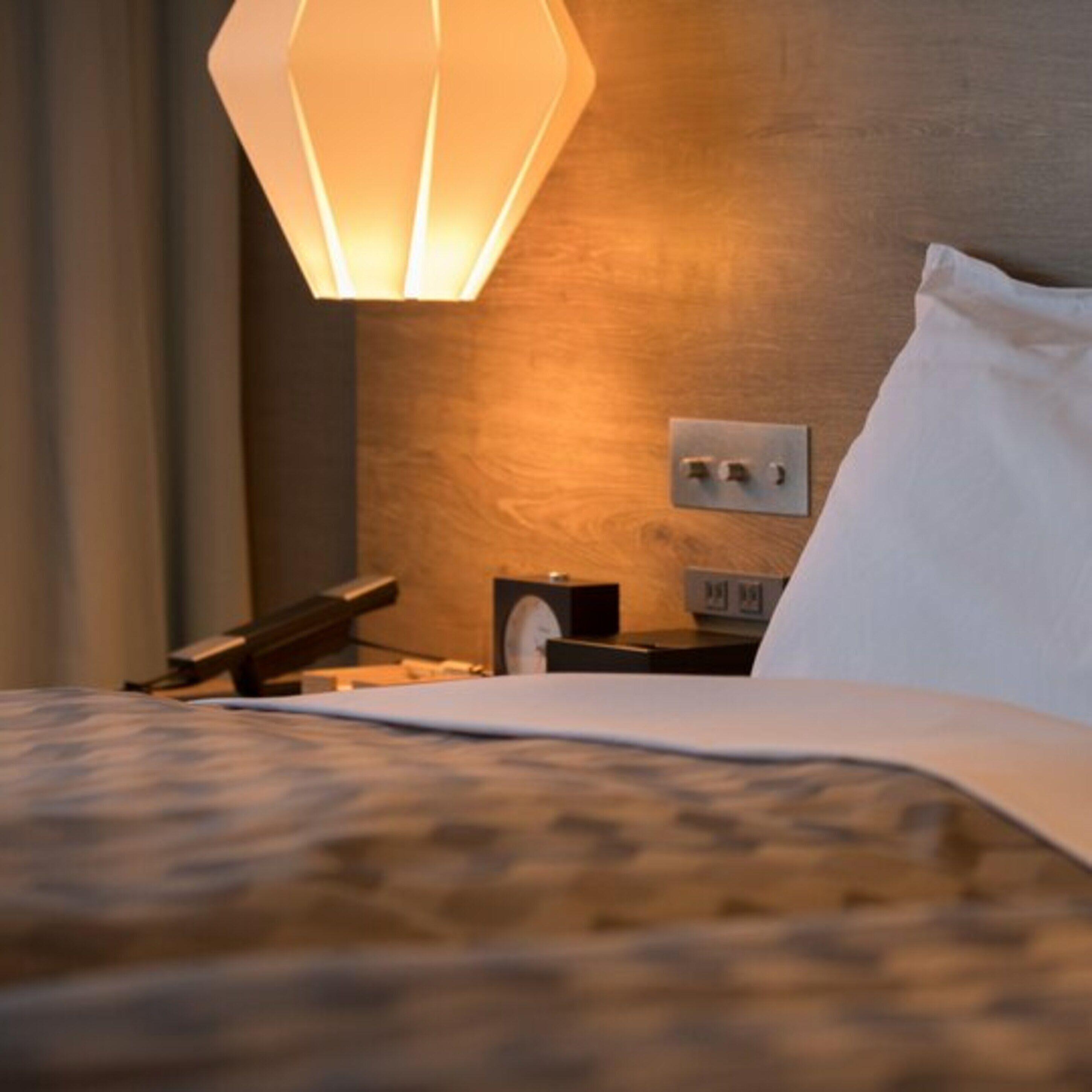 Una cama bien hecha con una colcha estampada, una lámpara de noche, una mesita de noche con un teléfono y un pequeño reloj, y un enchufe de pared.