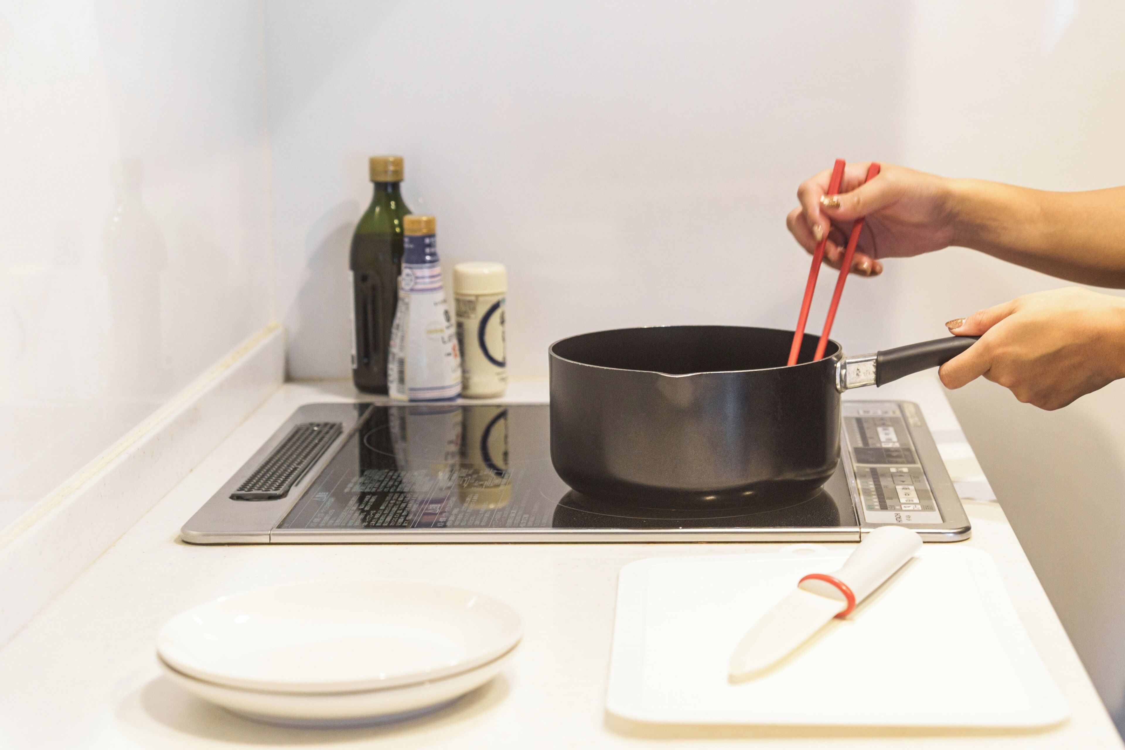 Une personne cuisine sur une plaque à induction, avec une casserole et des pinces rouges. On aperçoit en arrière-plan une assiette blanche, une planche à découper et des bouteilles de cuisine.