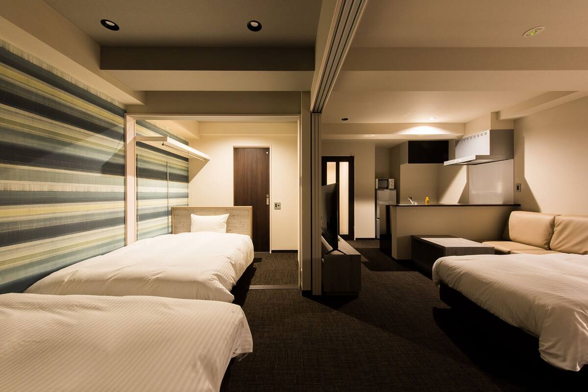 A hotel room with two beds, a sofa, a TV, and a kitchenette.