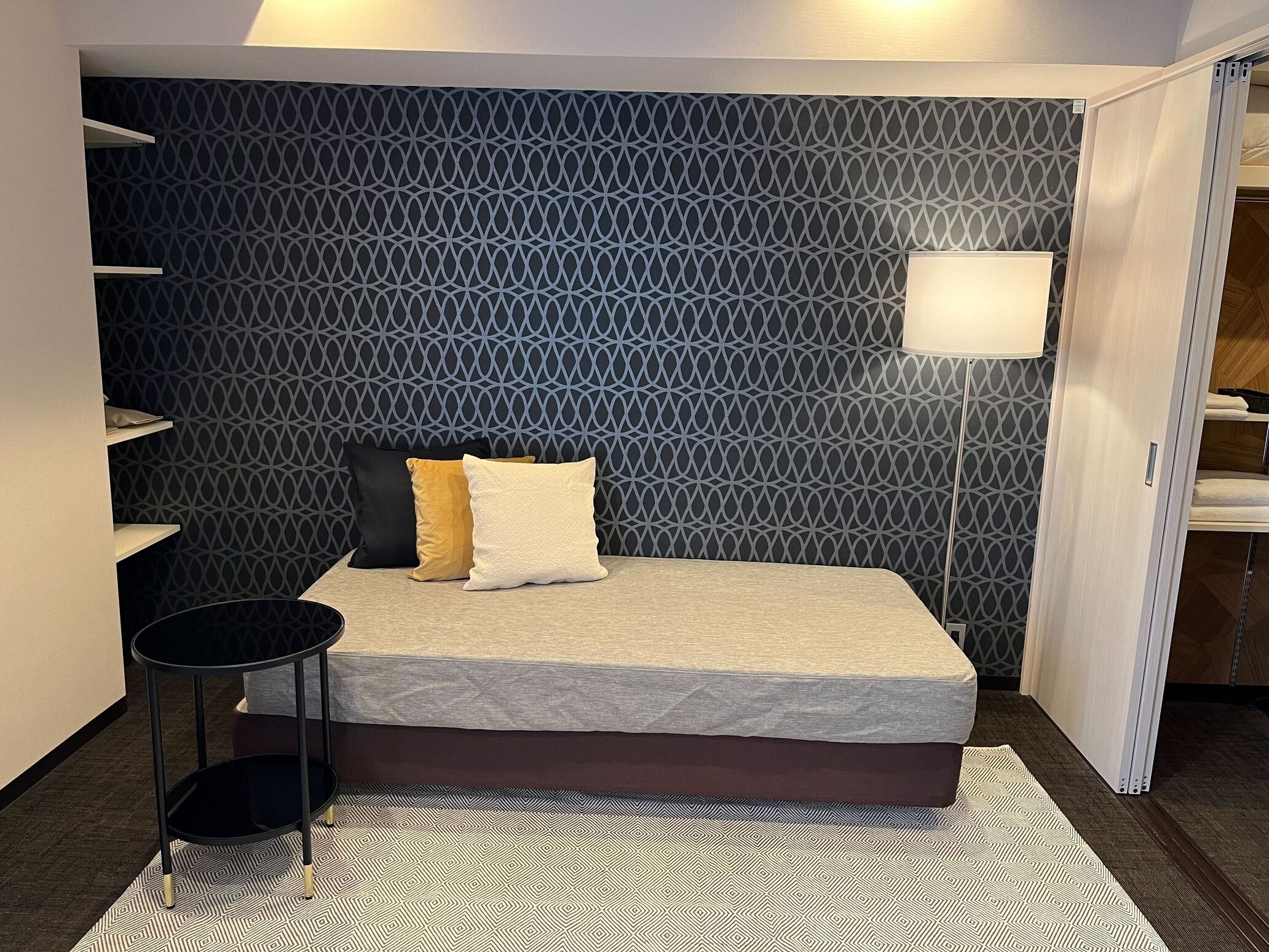 A compact bedroom with a patterned wall, a bed with two pillows, a bedside table, and a floor lamp.
