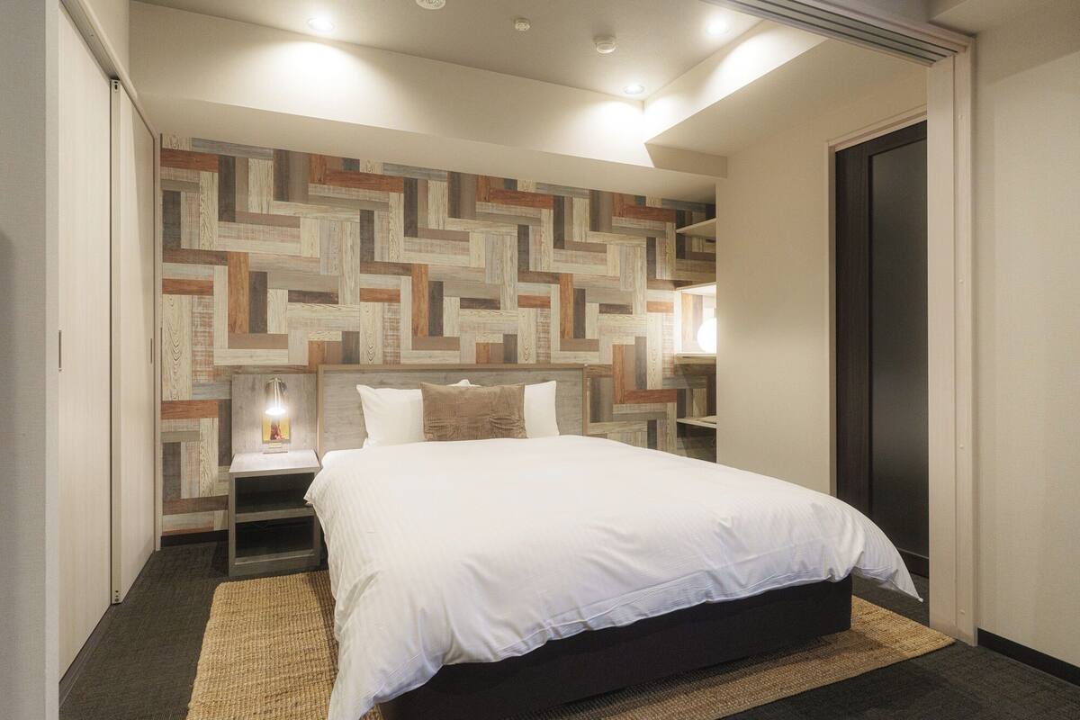 A modern hotel room with a large bed, a geometric-patterned headboard, a nightstand with a lamp, and a dark door.