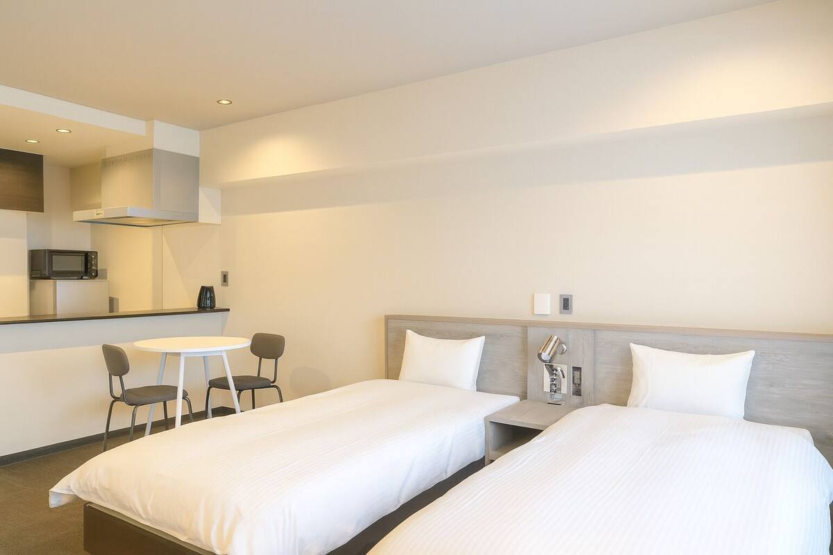 A hotel room with two single beds, a small kitchenette, a dining table with chairs, and a large mirror.