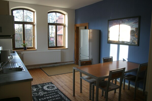 A kitchen with two windows, a refrigerator, a dining table with chairs, and a coffee-themed rug.