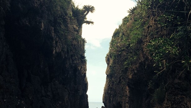 A narrow sandy path flanked by rocky cliffs with sparse vegetation, leading to a beach. - Mannnaya- (Motobu)