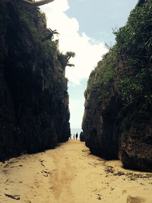 A narrow sandy path flanked by rocky cliffs with sparse vegetation, leading to a beach. - Mannnaya- (Motobu)