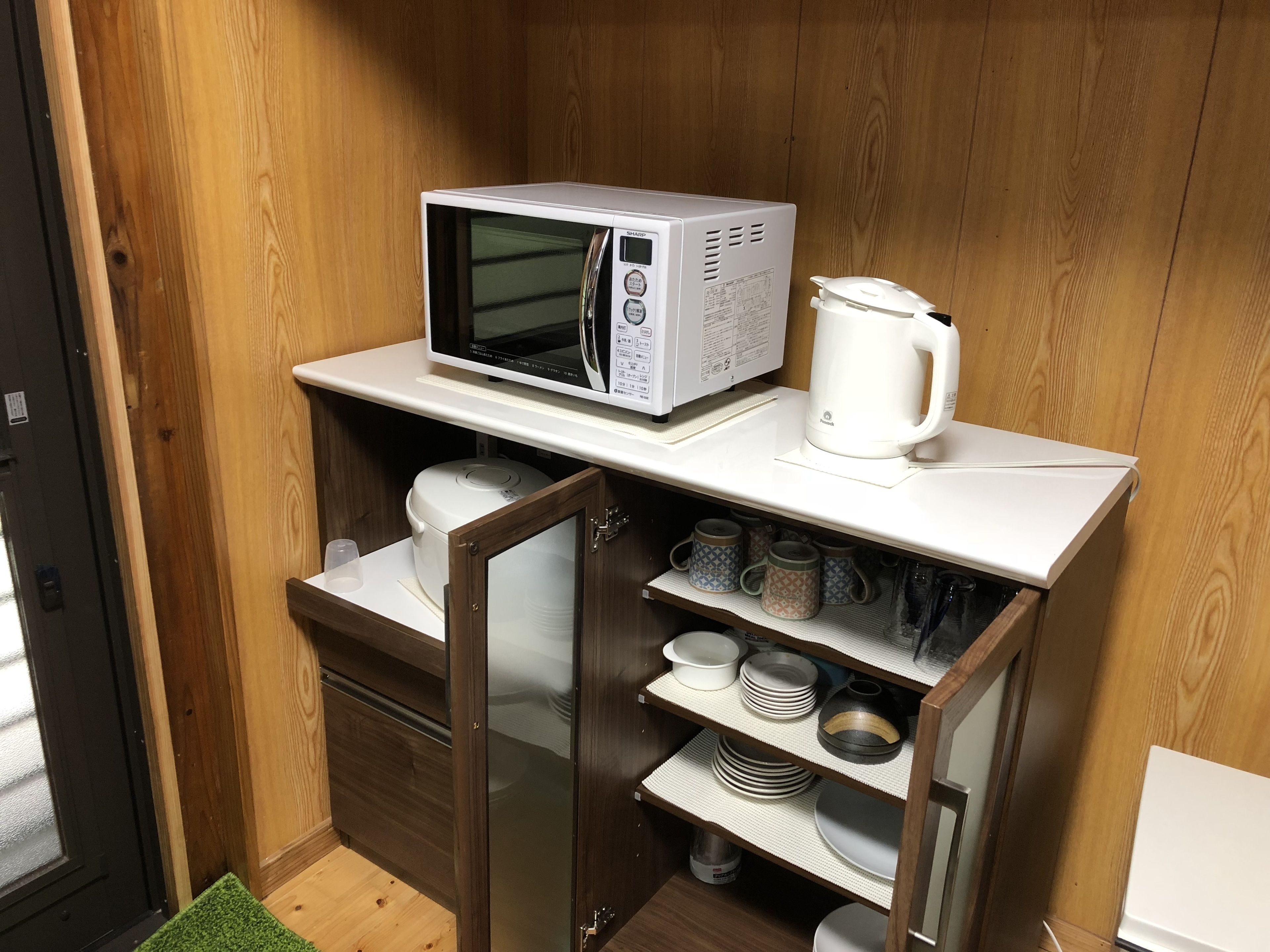 A compact kitchenette with a microwave, kettle, and shelves with dishes and cups.
