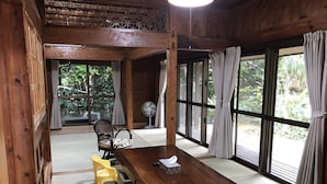 A wooden room with tatami flooring, a wooden table, a chair, a fan, and large windows with curtains. - Mannnaya- (Motobu)