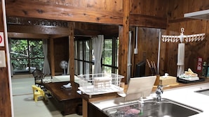 A wooden interior with a kitchen area including a sink, a dining table with chairs, and a view of greenery outside. - Mannnaya- (Motobu)