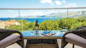 Ein Balkon mit Korbmöbeln, einer Flasche und Gläsern, mit Blick auf eine Küstenlandschaft mit Bäumen und Bergen.