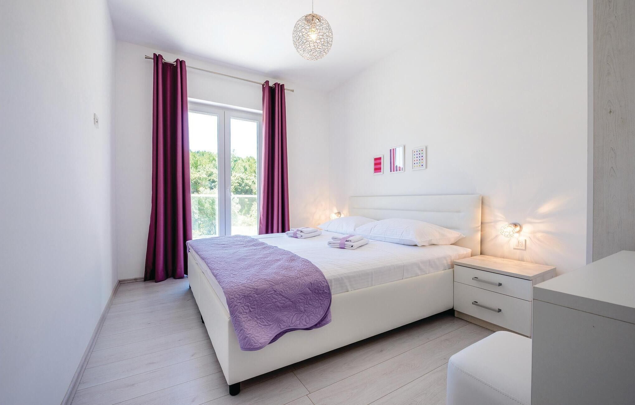 A bedroom with a bed, bedside tables, a nightstand, a window with curtains, and a view of greenery outside.