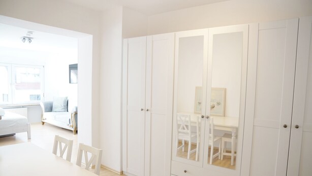 A bright, white room with a dining table and chairs, a large white wardrobe, and a glimpse of a living area with a sofa and a window. - F10 APARTMENT Ulm • Apartment in the heart of Ulm (Ulm)