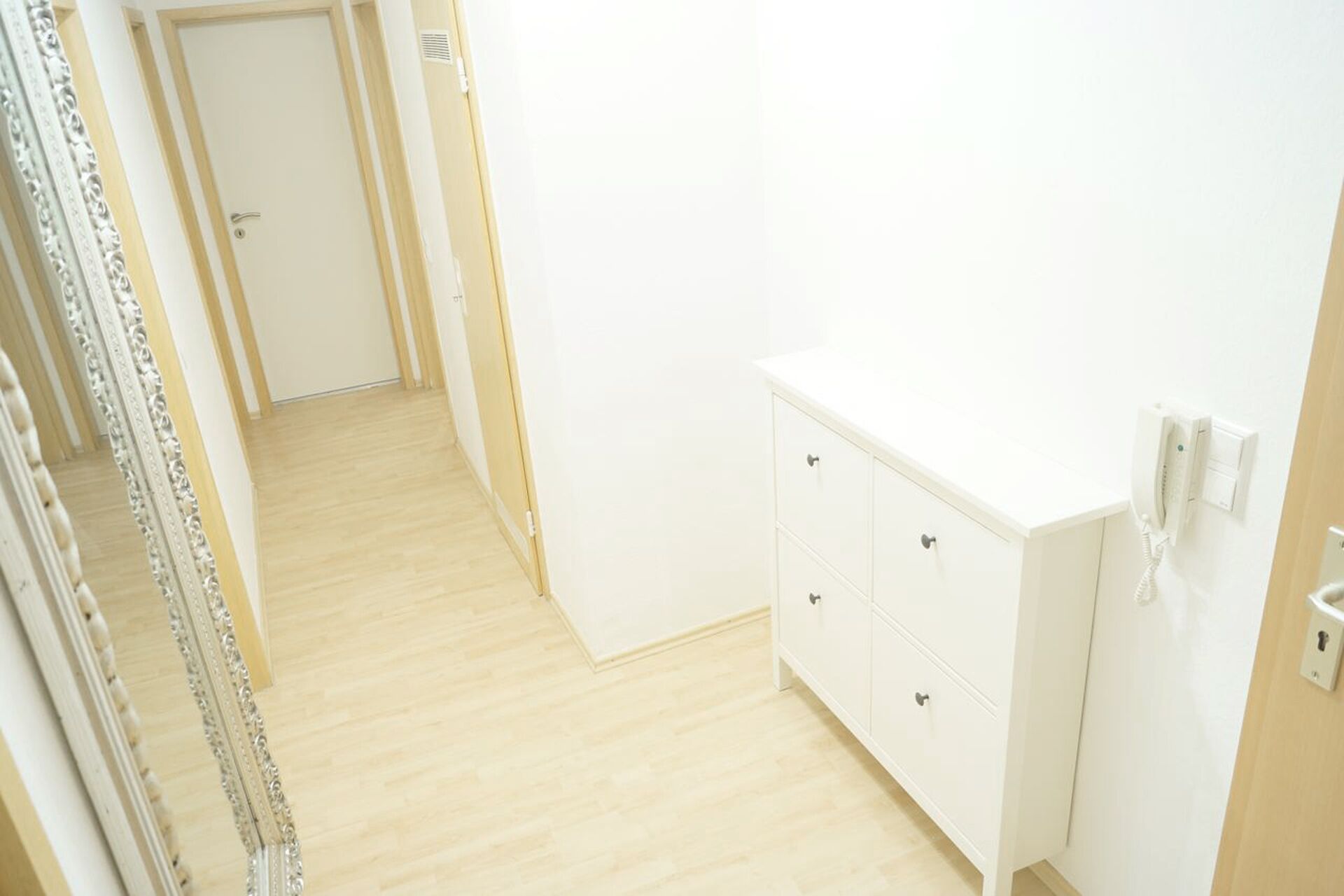 A narrow hallway with a wooden floor, a white door at the end, and a white cabinet with a telephone on the right.