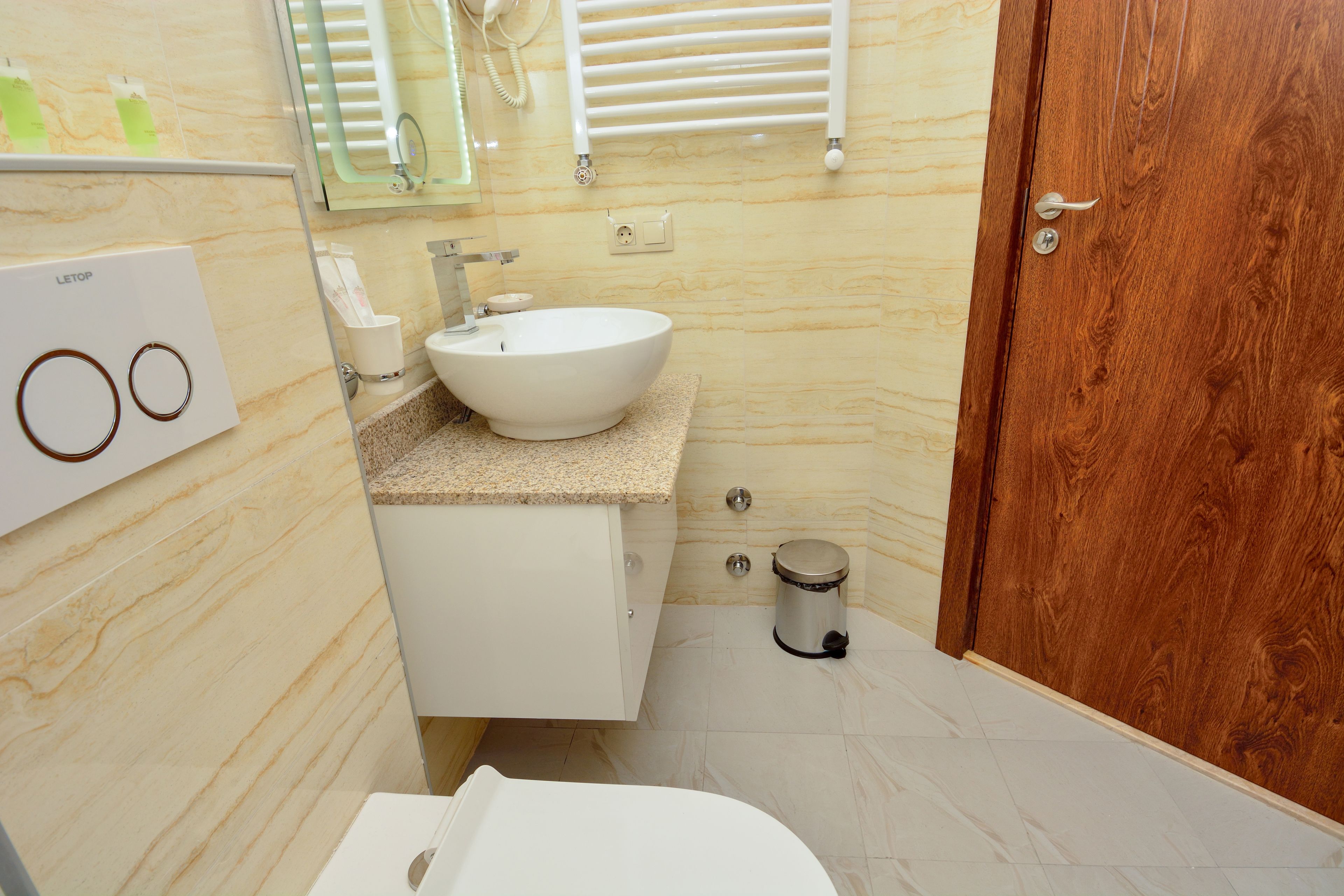 Un baño con lavamanos, tocador y retrete blancos. Se ve una puerta de madera y un cubo de basura.