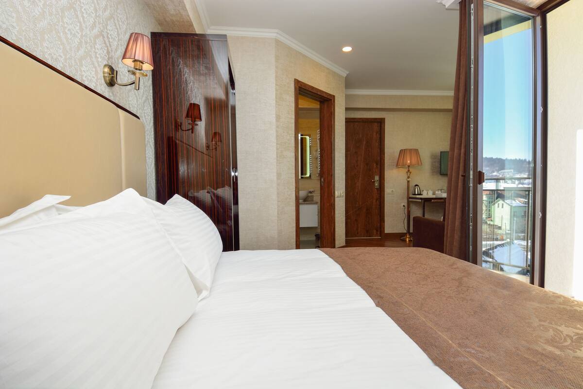 Une chambre d’hôtel avec un grand lit, une armoire en bois, une salle de bain avec un miroir et un balcon avec vue.