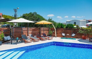 En swimmingpool med solstolar och parasoller, omgiven av ett trästaket och grönska.