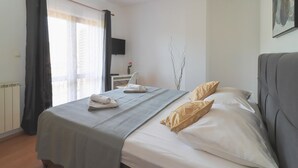 Een netjes opgemaakt bed met een grijze sprei en twee beige kussens, een klein nachtkastje met een opgevouwen handdoek, een wandtelevisie en een radiator.