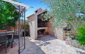 Une terrasse extérieure dotée d’un espace barbecue en pierre, d’une pergola en métal et d’un coin salon avec des chaises en métal.