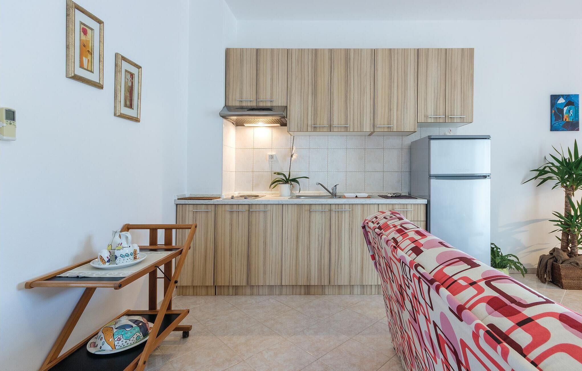 A compact kitchen with wooden cabinets, a built-in stove, and a refrigerator. There is a small dining table with a chair, a potted plant, and a colorful cushion.