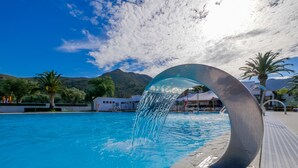 Une piscine agrémentée d’une fontaine, entourée de palmiers et de bâtiments, sous un ciel dégagé.