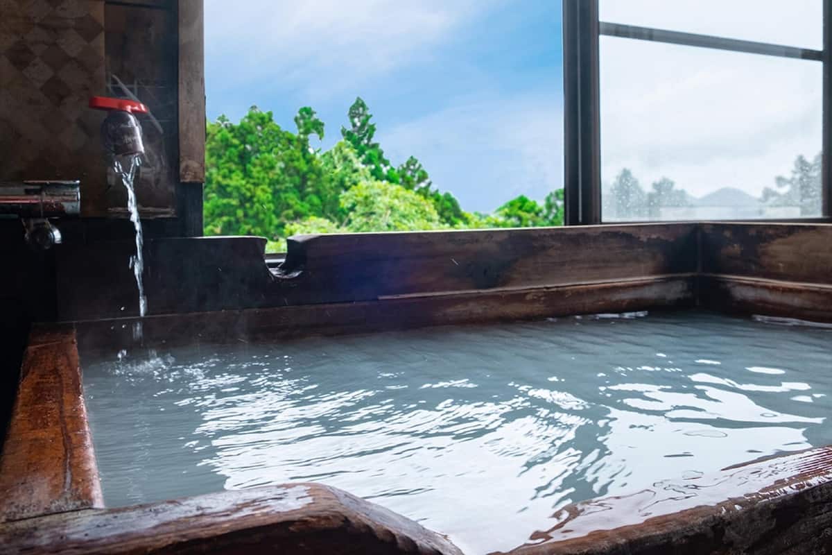 Une baignoire en bois avec un robinet, une fenêtre donnant sur un paysage verdoyant et un mur en pierre.