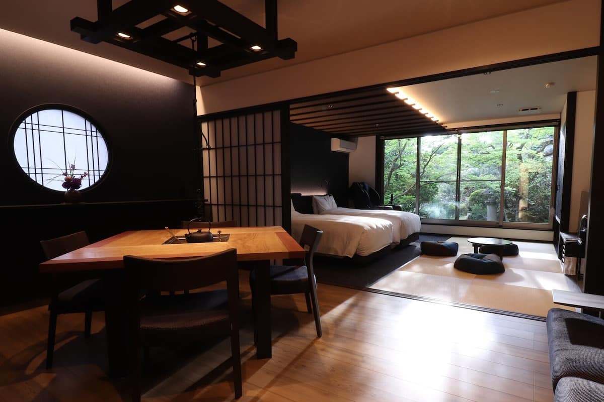 Una habitación tradicional japonesa con suelo de tatami, una mesa baja con sillas, una cama con futón y un ventanal con vistas a zonas verdes.