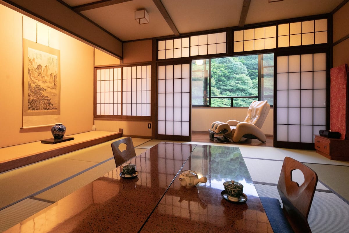 Una habitación japonesa tradicional con suelo de tatami, una mesa baja, una silla acolchada, un juego de té y un cuadro en la pared.