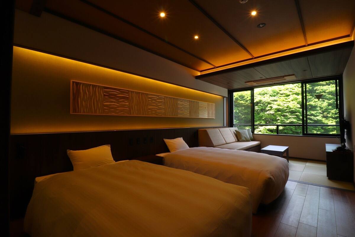 A hotel room with two beds, a large window with a view of greenery, and a wall-mounted artwork.