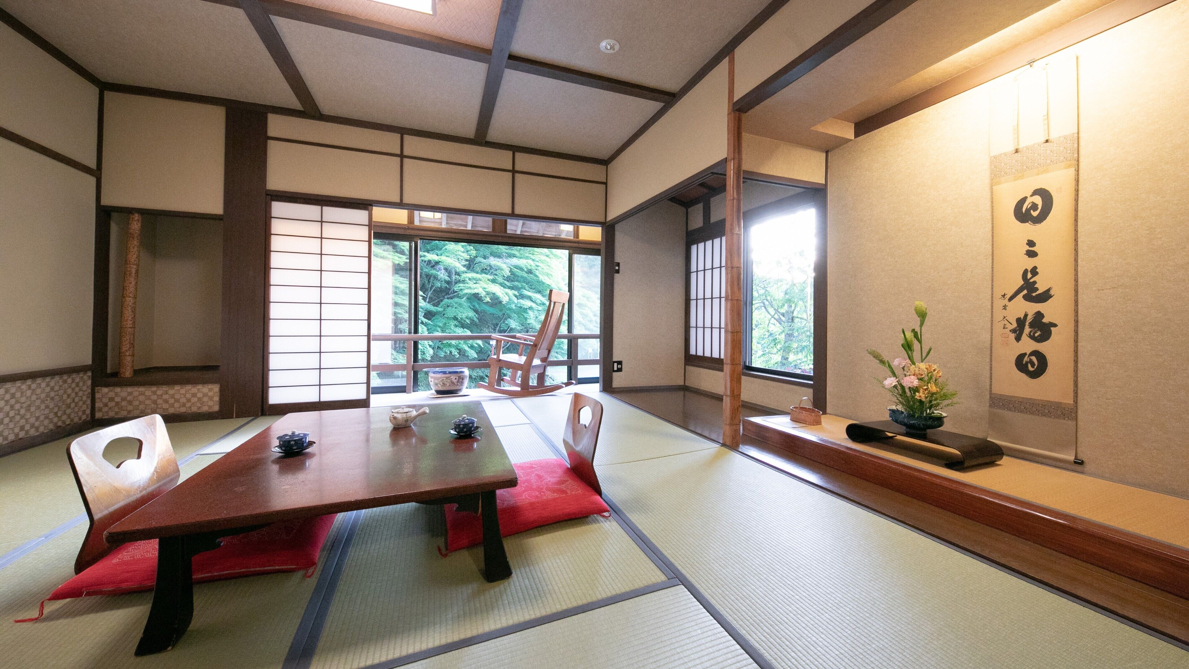 A traditional Japanese room with tatami flooring, a low table, a tea set, a rocking chair, and a view of trees through large windows.