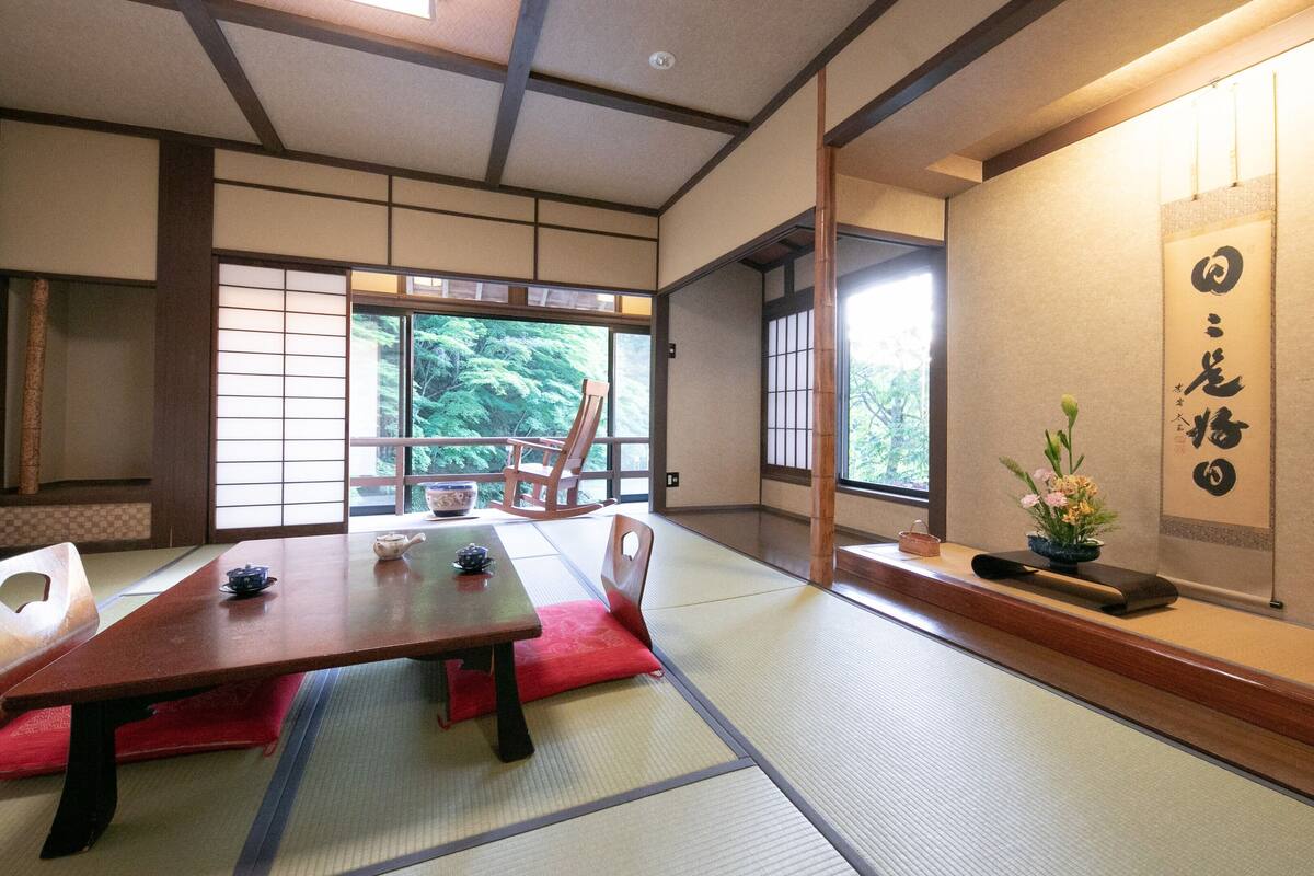 Una habitación japonesa tradicional con suelo de tatami, una mesa baja, un juego de té, un sillón mecedora y vistas a árboles a través de amplias ventanas.