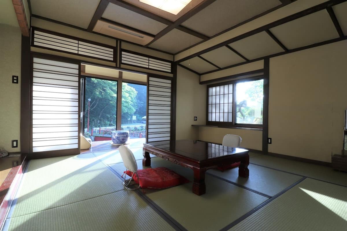 Una habitación japonesa tradicional con suelo de tatami, una mesa de madera baja y puertas correderas que dan a vistas de vegetación.