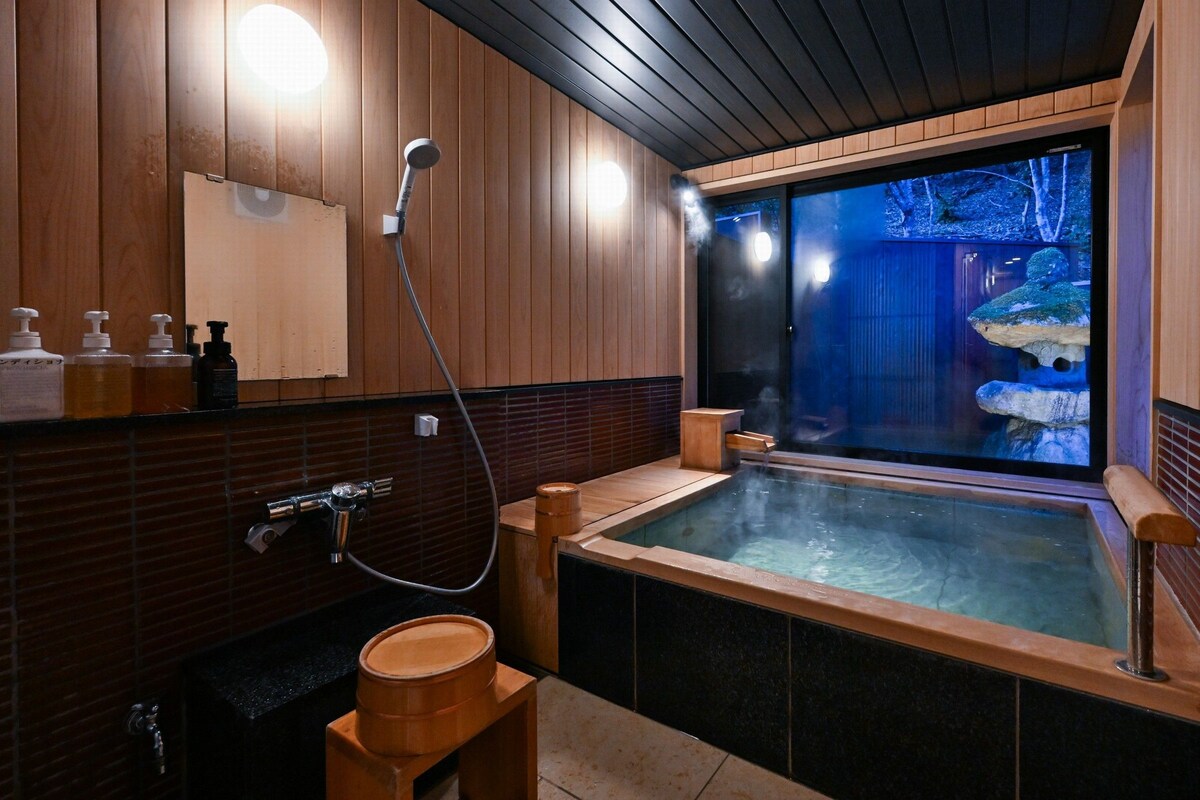 Ein modernes Badezimmer mit Holzvertäfelung, einer großen Badewanne, einem Spiegel und Blick auf einen Wald durch eine Glastür.