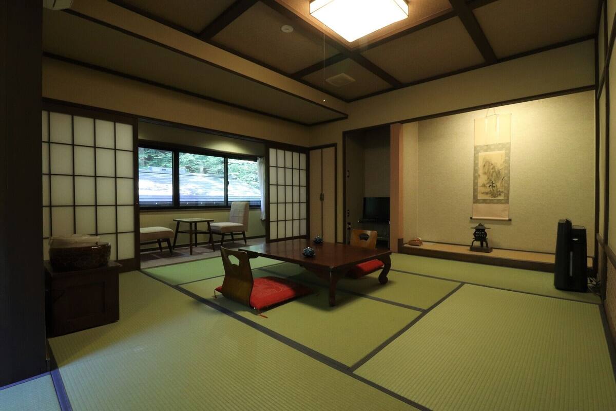 Una habitación japonesa tradicional con suelo de tatami, una mesa baja, sillas, un televisor y una puerta corredera.