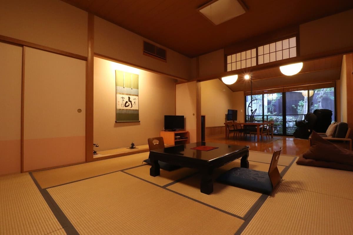 Una habitación japonesa tradicional con suelo de tatami, una mesa baja, un televisor y vistas a un jardín a través de amplios ventanales.